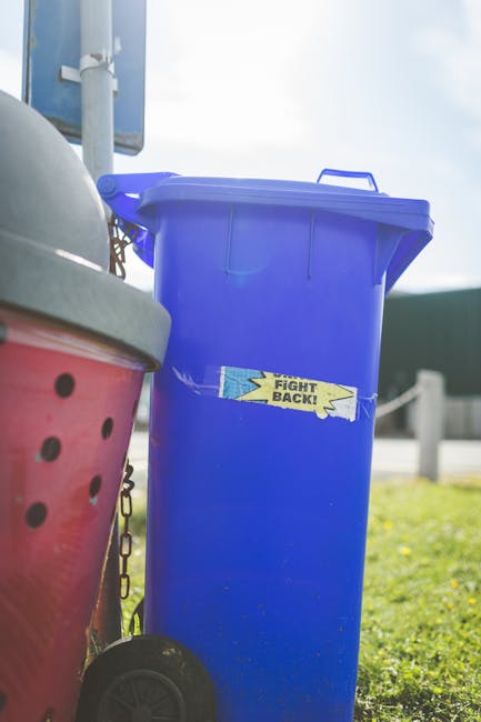 A tall, rectangular blue waste bin with a closed lid, positioned outdoors on a grassy area. The bin has a smooth, plastic surface with a faint sheen and a yellow and blue sticker on its front that reads 'FIGHT BACK!'. To the left of the blue bin, part of a red container with small round ventilation holes and a slightly curved surface is visible, both sitting on a pavement. Behind these bins, there is a white pole with a mounted sign and a partially visible blue structure or sign. The background includes a bright, partly cloudy sky and some green (possibly a hedge or fence), indicating natural daylight. The scene suggests a setup for an outdoor waste management or recycling collection point, likely related to private or alternative waste handling services such as those offered by waste removal specialists like Waste Removal Richmond. The environment appears organized, with a focus on on-site rubbish storage and disposal.