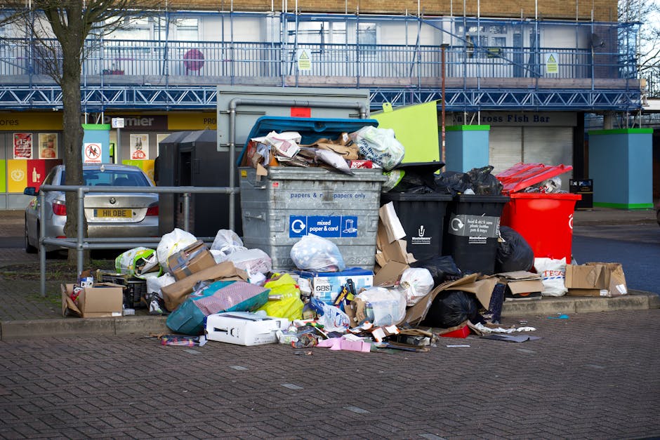 A collection of overflowing rubbish bins and scattered trash bags, cardboard boxes, paper, plastic packaging, and miscellaneous waste items, situated on a paved urban street corner in front of a commercial building with a shopfront and signage. The waste includes a large grey mixed paper and cardboard recycling bin with its lid partially open, revealing a jumble of paper and packaging materials inside, alongside black and red bins for general waste. Several black and plastic trash bags are piled around the bins, some torn open with loose waste spilling onto the ground. Additional smaller packaging boxes, plastic containers, and discarded paper items are strewn across the pavement. In the background, a parked silver car is visible behind a metal railing, and the building behind features blue scaffolding or netting, with storefront signage indicating local shops. The scene suggests a situation involving private rubbish collection, typical of an independent waste disposal service, demonstrating the importance of timely rubbish removal and clearance in urban environments.