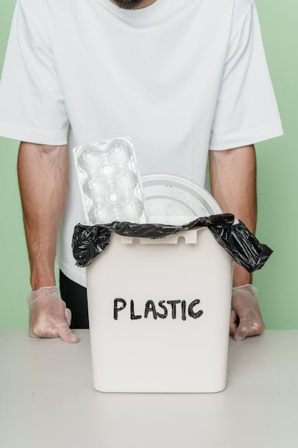A person wearing a white T-shirt and transparent disposable gloves stands behind a white plastic bin labeled 'PLASTIC' in black, handwritten style. The bin's lid is partially open, with a black plastic trash bag tucked inside, visible around the edges. Inside the bin, there is an empty clear plastic egg carton positioned upright and partially protruding from the opening. The background features a plain, light green wall, and the scene is lit with even lighting, emphasizing the contents and the person's hands. The image visually relates to waste management and the collection of plastic waste, aligning with services for rubbish removal and environmentally responsible disposal, as offered by Waste Removal Richmond.